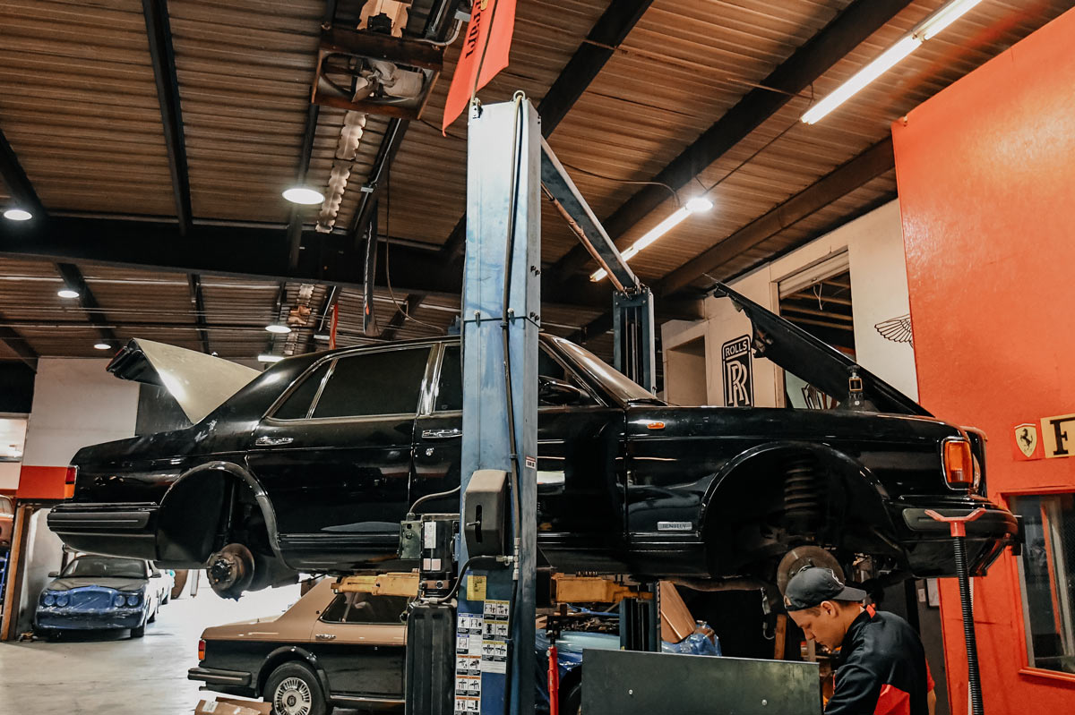 Classic Bentley elevated on lift with technician working below at European Auto Service shop in Reseda with Rolls-Royce and Ferrari branding on the wall