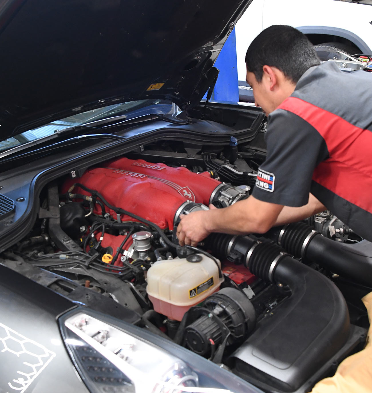 Technician inspecting Ferrari engine bay with red intake manifold and coolant reservoir during diagnostic evaluation at European Auto Service