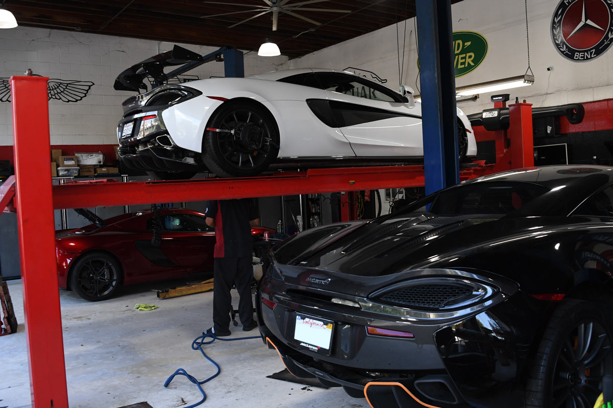 Three McLarens being serviced simultaneously including white 570S on lift and black and red models on the floor at European Auto Service with Aston Martin and Land Rover logos