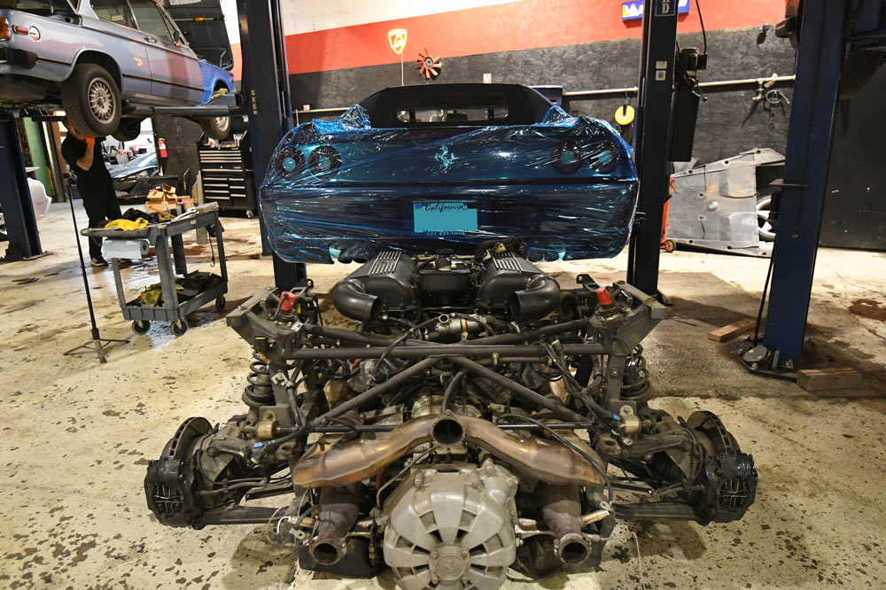 Ferrari F355 V8 engine and transaxle assembly on shop floor after removal, car raised on lift behind it — European Auto Service Los Angeles